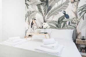 A neatly made bed with white linens and towels, a bedside table with a tray holding a cup and a book, and a wall with a tropical mural featuring birds and foliage. - Easylife - Apartment with terrace near Cenacolo (Milano)