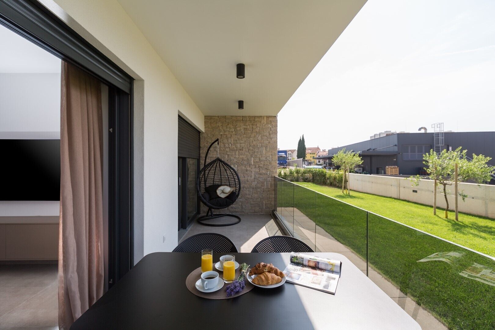 A balcony with a hanging chair, a table with breakfast, and a view of a green lawn and buildings.