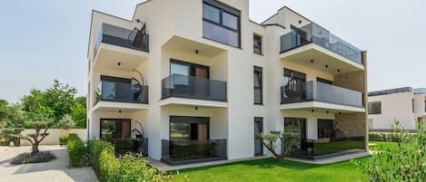 A modern multi-story residential building with balconies, a well-maintained lawn, and a clear sky.