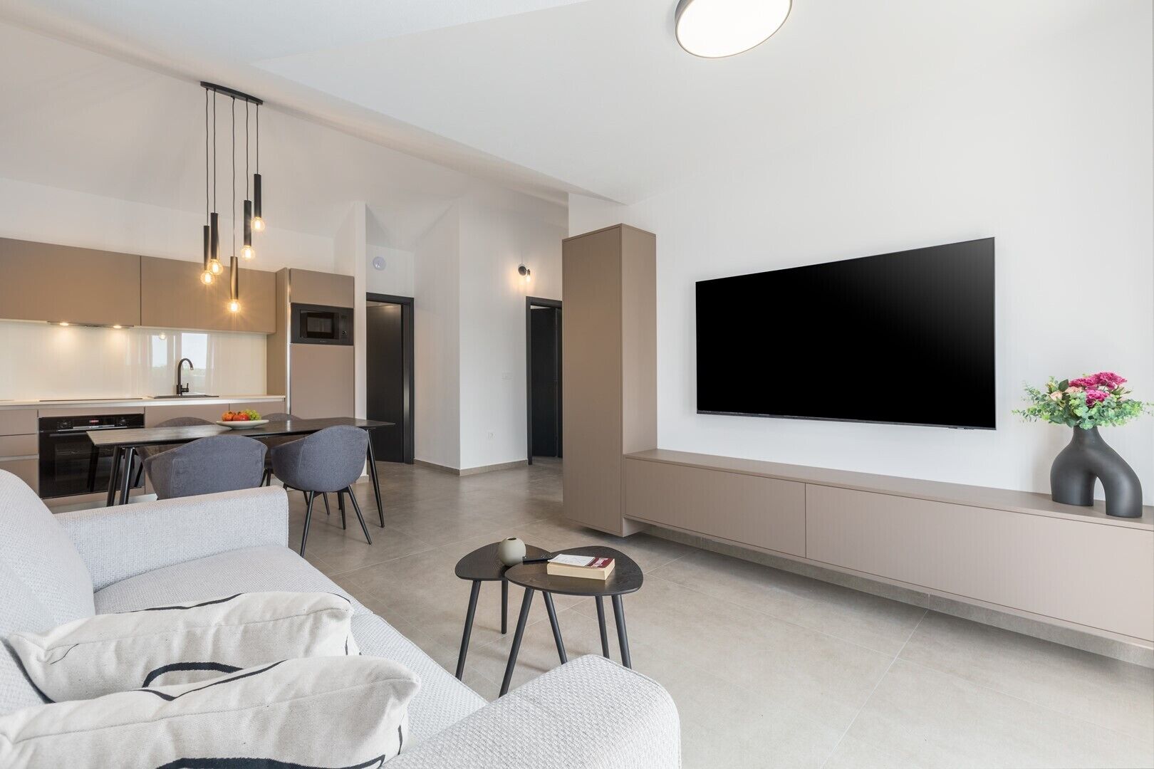 Ein modernes Wohnzimmer mit einem großen Flachbildfernseher an der Wand, einer Couch, einem Couchtisch und einem Essbereich mit Stühlen. Im Hintergrund befindet sich eine Küche.