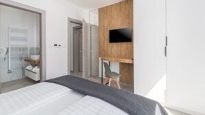 Une chambre moderne avec un lit, un téléviseur fixé sur un panneau en bois, une chaise et une salle de bain équipée d’un porte-serviettes et d’un lavabo.