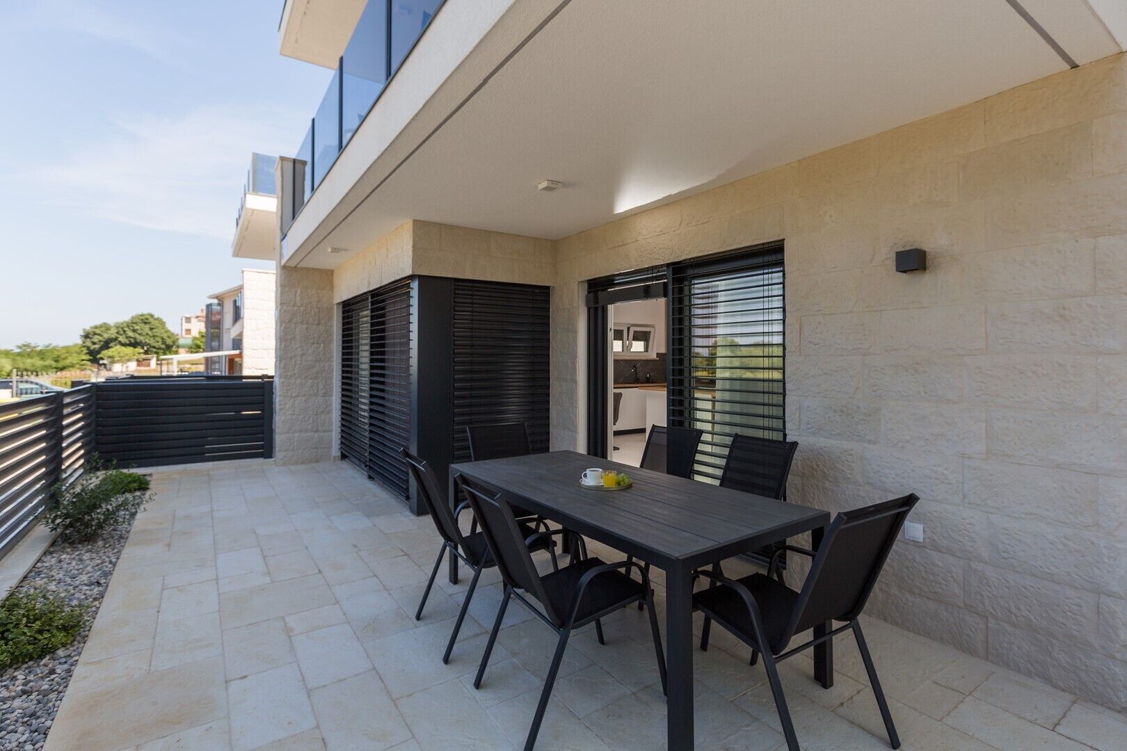 A modern outdoor patio with a dining table and chairs, a kitchen area visible through a window, and a stone wall.