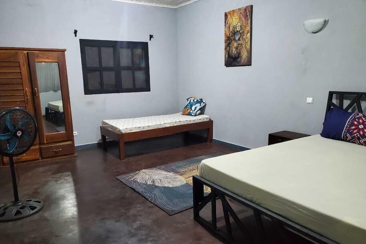 Une chambre à coucher avec un lit, un banc, un ventilateur, une fenêtre et un tableau accroché au mur.