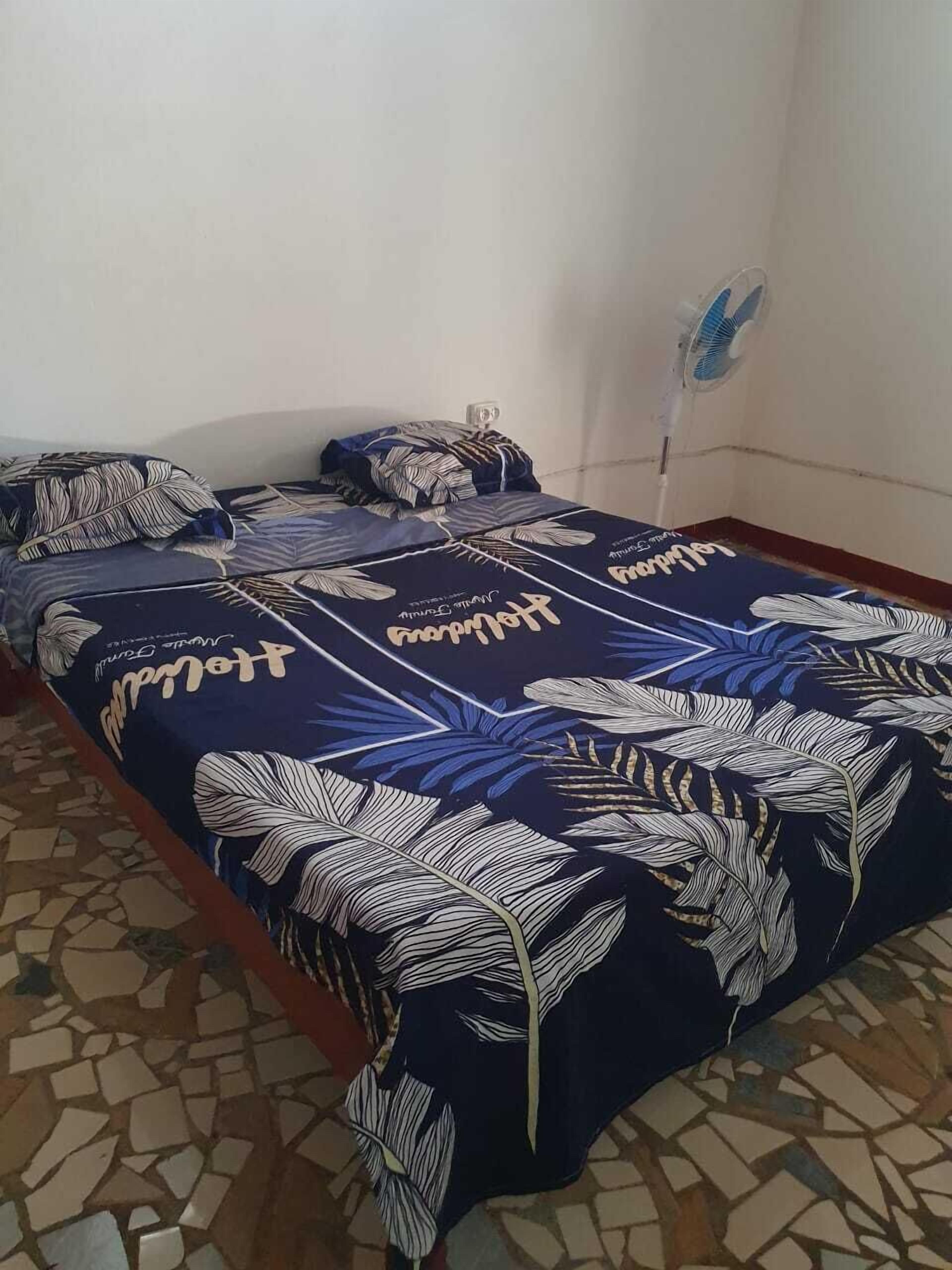 A bed with a floral bedspread, a wall-mounted fan, and a tiled floor.