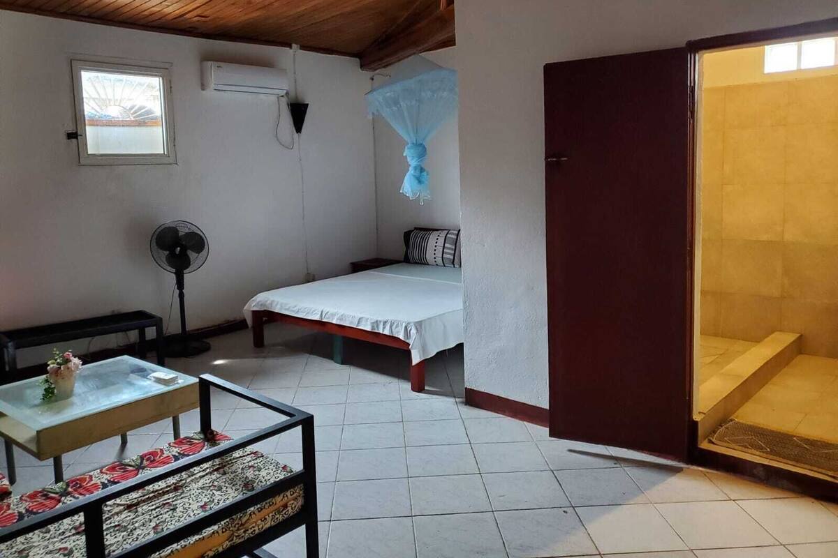 Une pièce avec un lit, une petite table sur laquelle se trouve un vase, un ventilateur et une fenêtre.