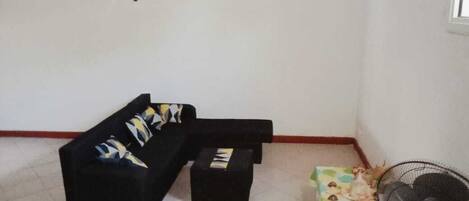A living room with a black sofa, a small coffee table, a colorful ottoman, and a standing fan.