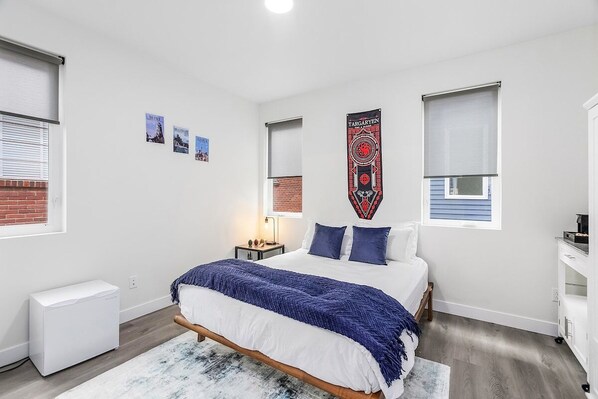Une chambre à coucher moderne, avec un lit, une petite table de chevet, une œuvre d’art accrochée au mur et une fenêtre avec des stores.
