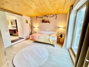 A bedroom with a wooden ceiling, a bed with a white headboard, a bedside table with a lamp, and a window with curtains.