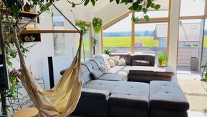 Una sala de estar moderna con un amplio sofá modular, una hamaca colgante y vistas a un campo verde a través de amplios ventanales.