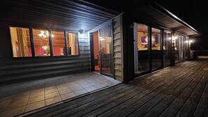 En modern byggnad med ett trädäck, stora glasdörrar och en inre belysning som syns genom fönstren.