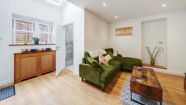 Ein Wohnzimmer mit einem grünen Sofa, einem Holztisch und einer Holzkommode mit Flechtfront. Ein Fenster mit Jalousien und eine Tür mit Glasfenster sind ebenfalls vorhanden.