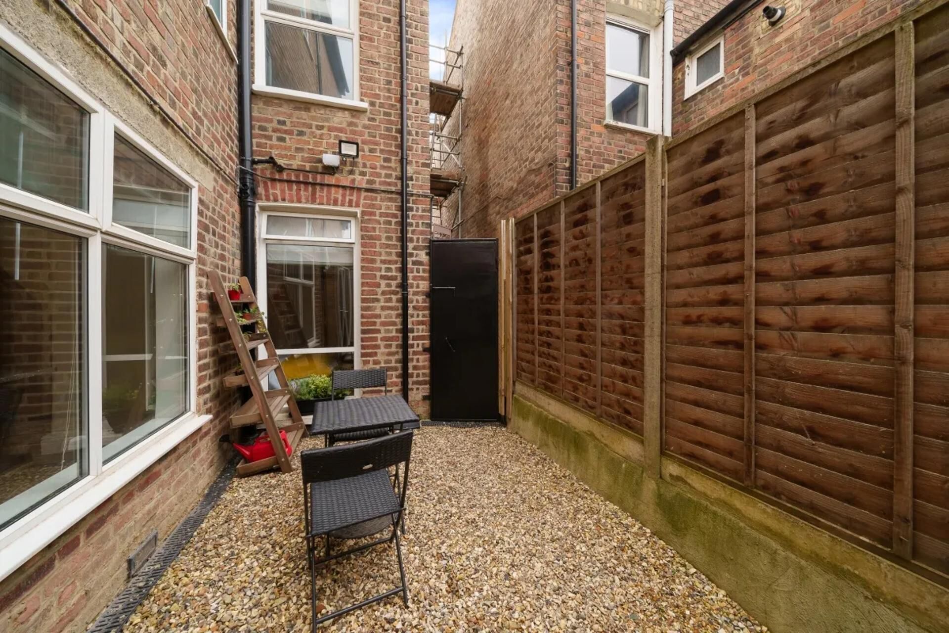 A narrow alleyway with a gravel path, a black metal chair, a ladder leaning against a brick wall, and a black door.