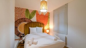 Un lit bien fait, avec du linge de lit blanc et une serviette enroulée, une lampe fixée au mur et une fenêtre avec des stores.