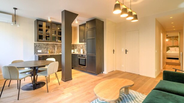 Une cuisine moderne avec des meubles de couleur sombre, un coin repas avec une table ronde et des chaises, et un salon accueillant avec un canapé vert et une petite table en bois.