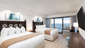 Ett hotellrum med två sängar, ett skrivbord, en TV och en balkong med utsikt över havet.