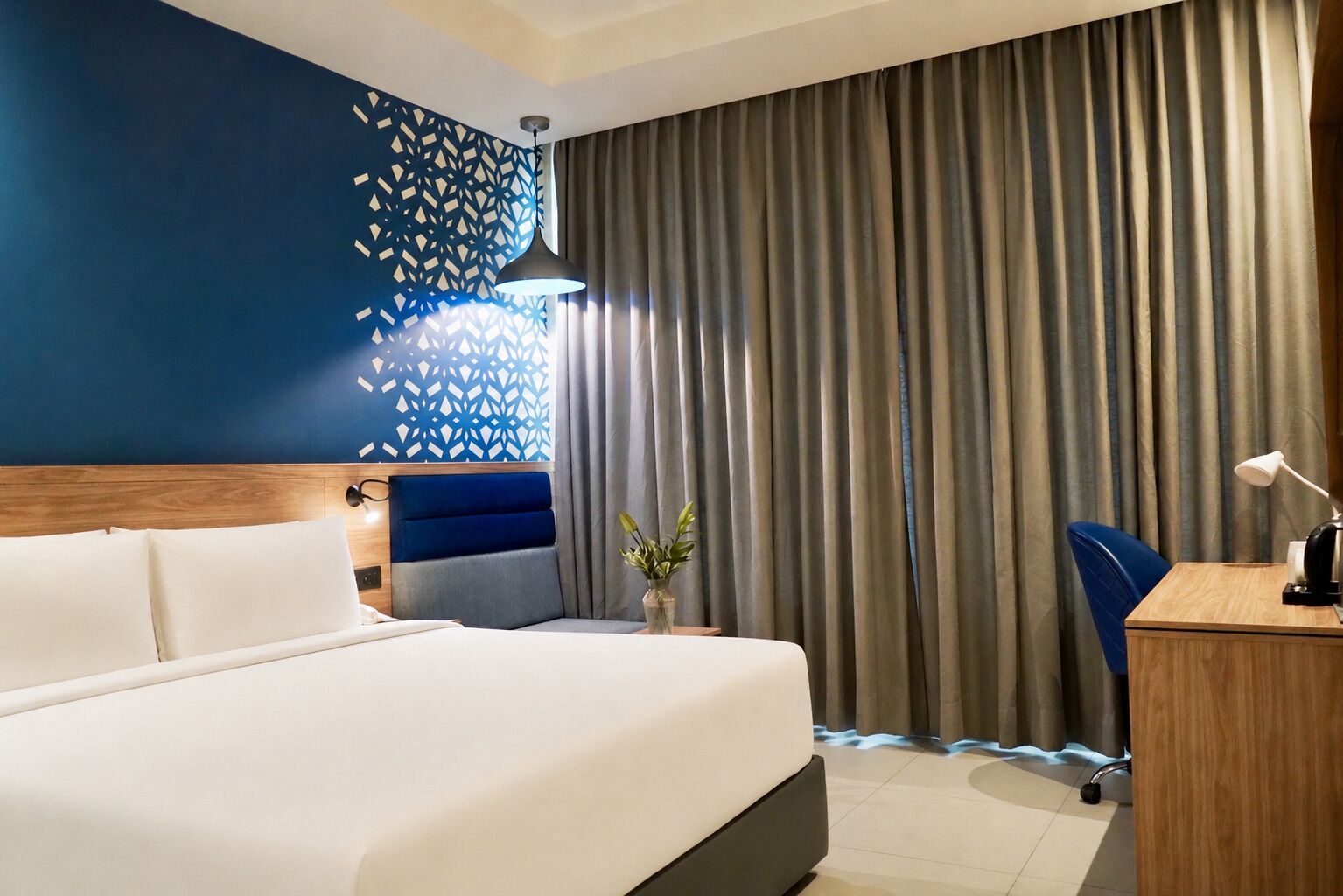 A hotel room with a bed, a desk, a chair, a vase with plants, and a decorative wall panel.