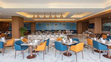 A modern restaurant with a central buffet, blue and orange chairs, and a wooden ceiling.