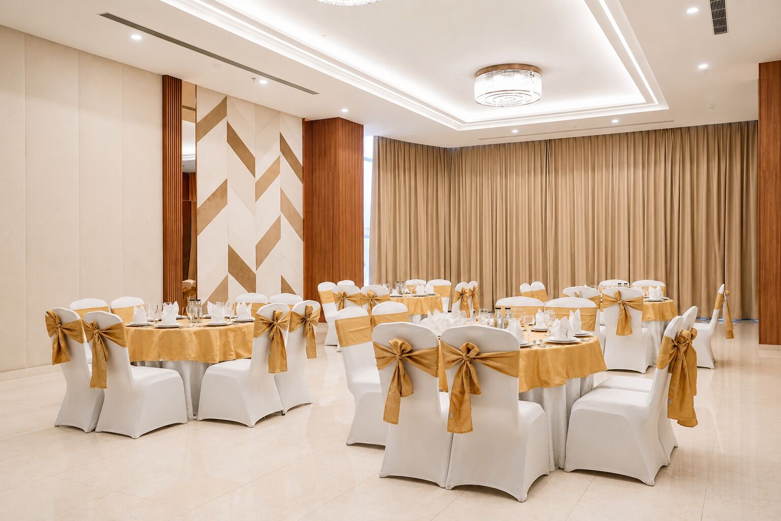 A banquet hall with round tables, white chairs with gold sashes, and a modern chandelier.