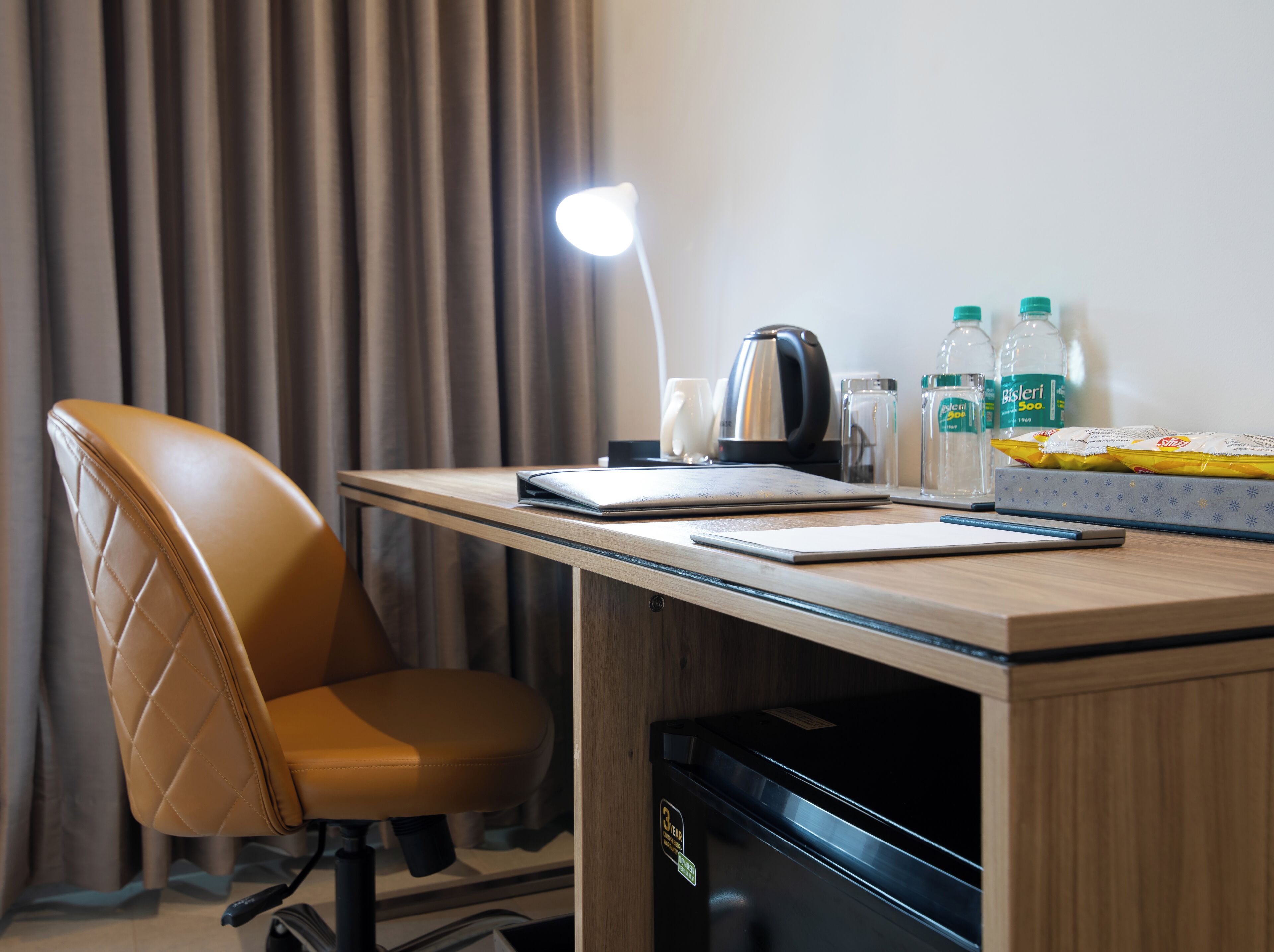Et hotelværelse med skrivebord, stol, lampe, elkedel, vandflasker og snacks.