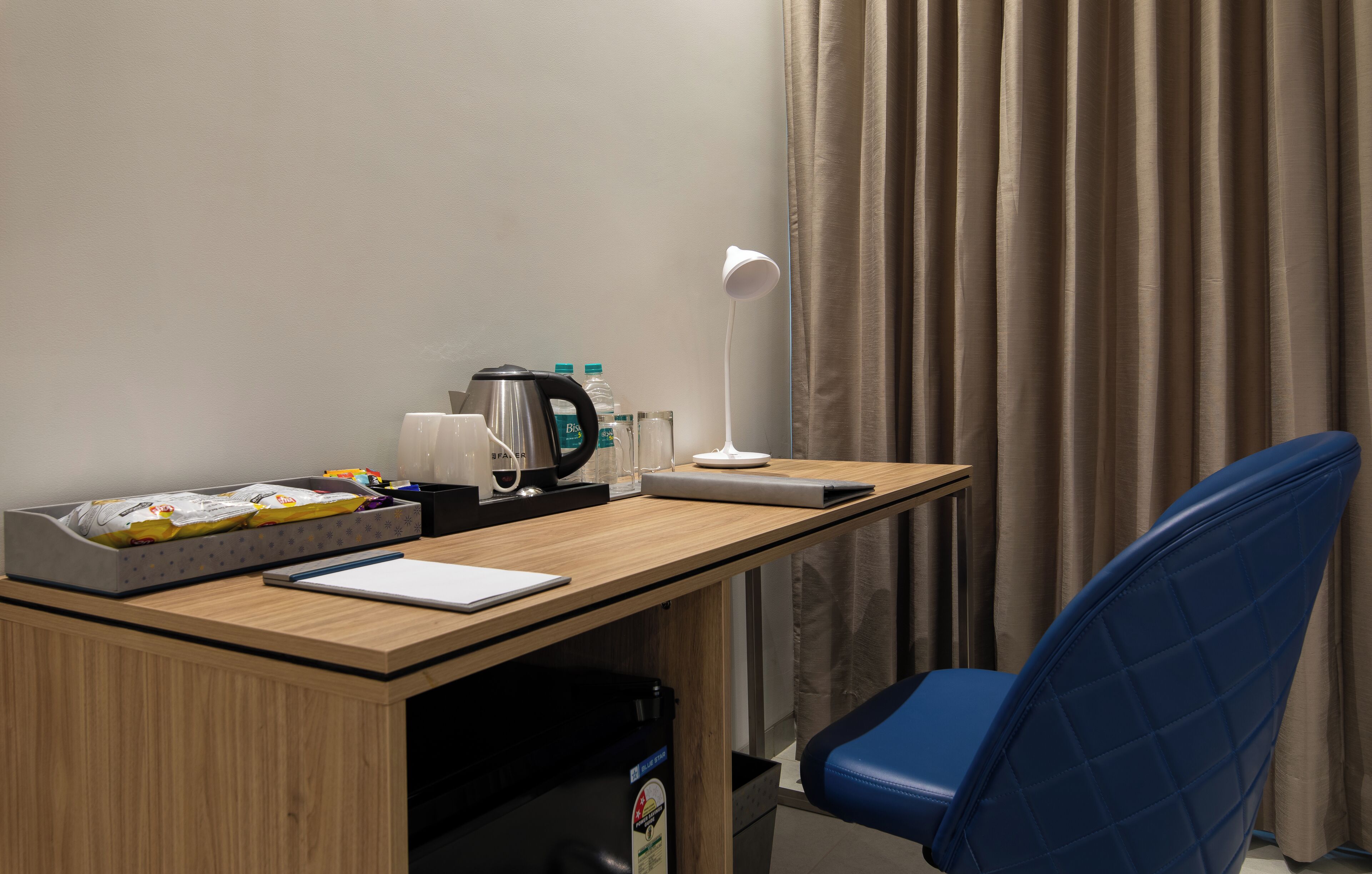 A hotel room with a wooden desk, a blue chair, a white desk lamp, a coffee maker, and a bottle of water.