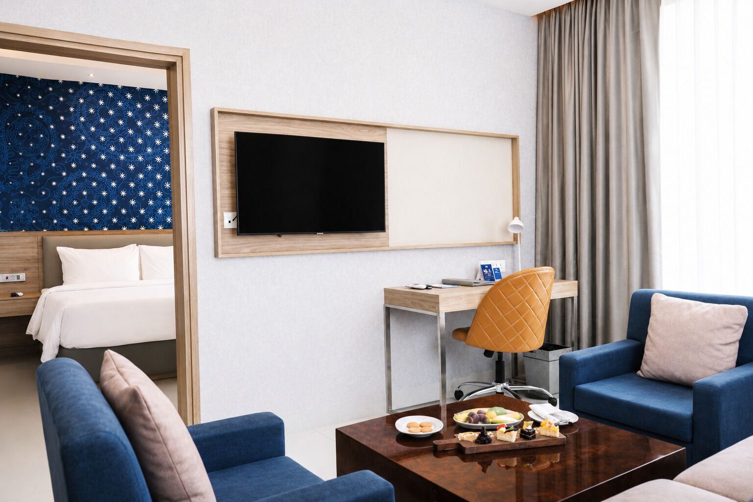 A hotel room with a bed, a desk with a chair, a TV, and a coffee table with snacks.