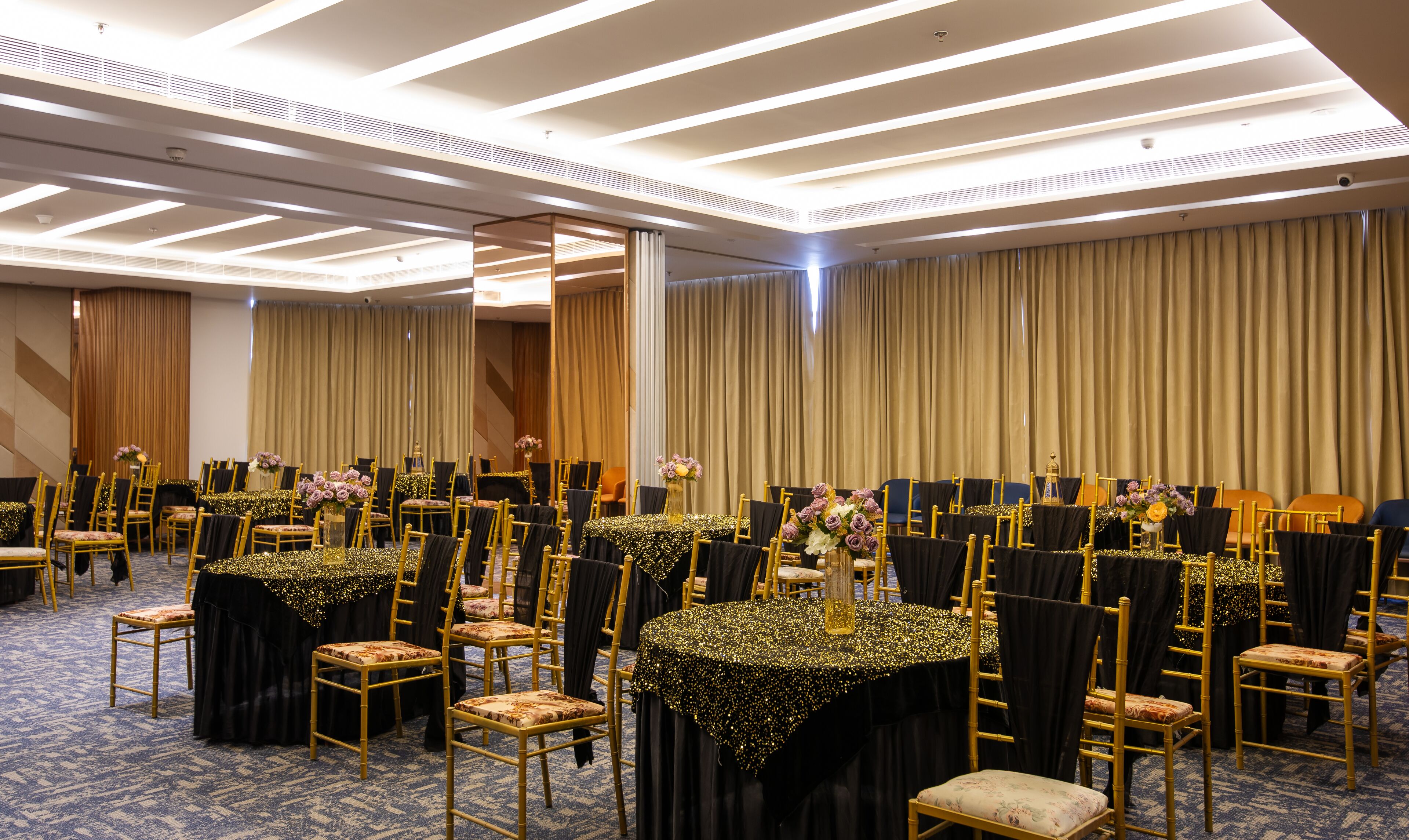 A banquet hall with rows of tables and chairs, a high ceiling with linear lighting, and a large curtain backdrop.