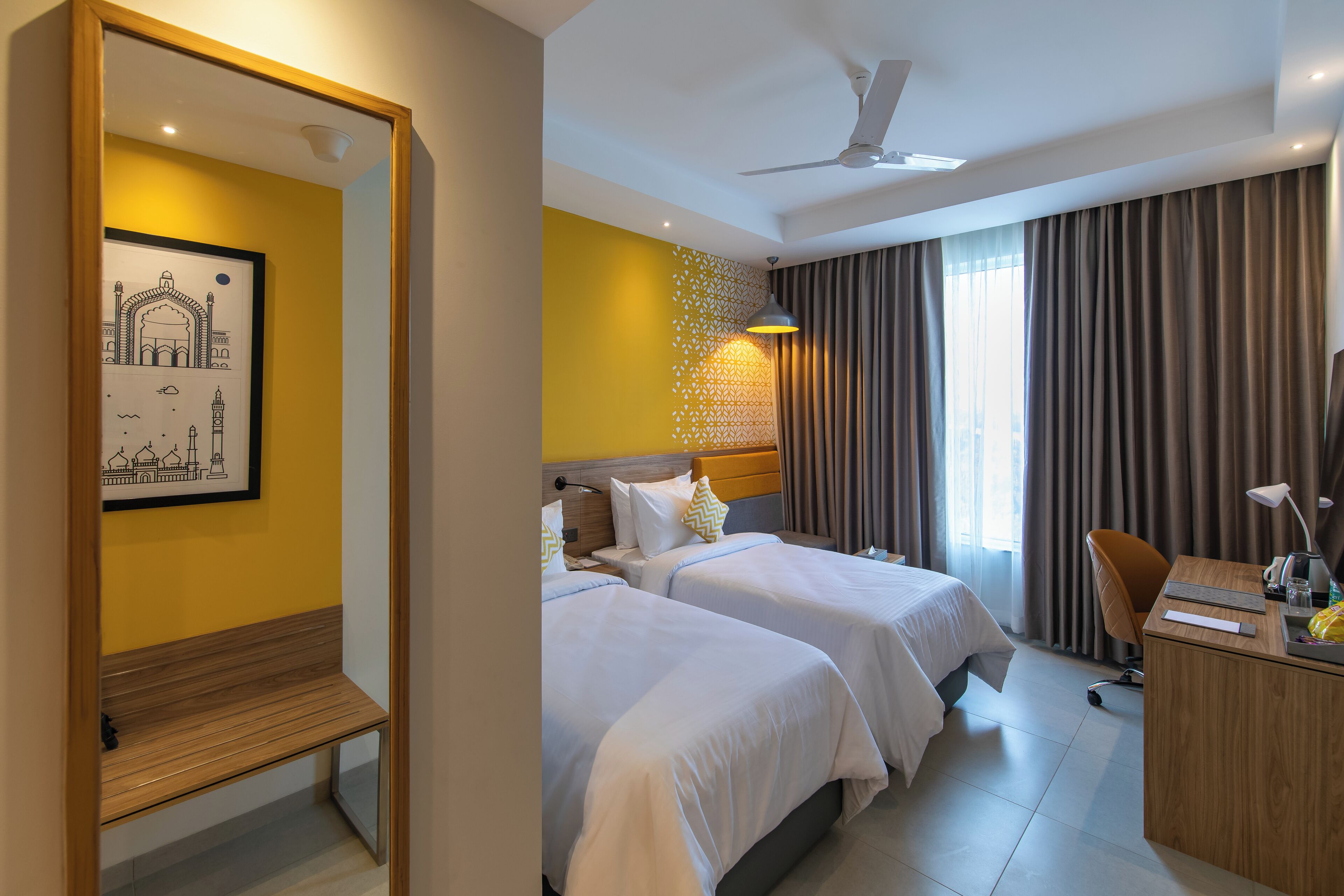 A hotel room with two beds, a desk, a chair, a ceiling fan, and a large mirror.