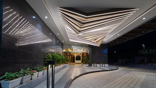 Ingresso moderno di un edificio con un'installazione luminosa geometrica al soffitto, un vialetto curvo e piante in vaso lungo il percorso.