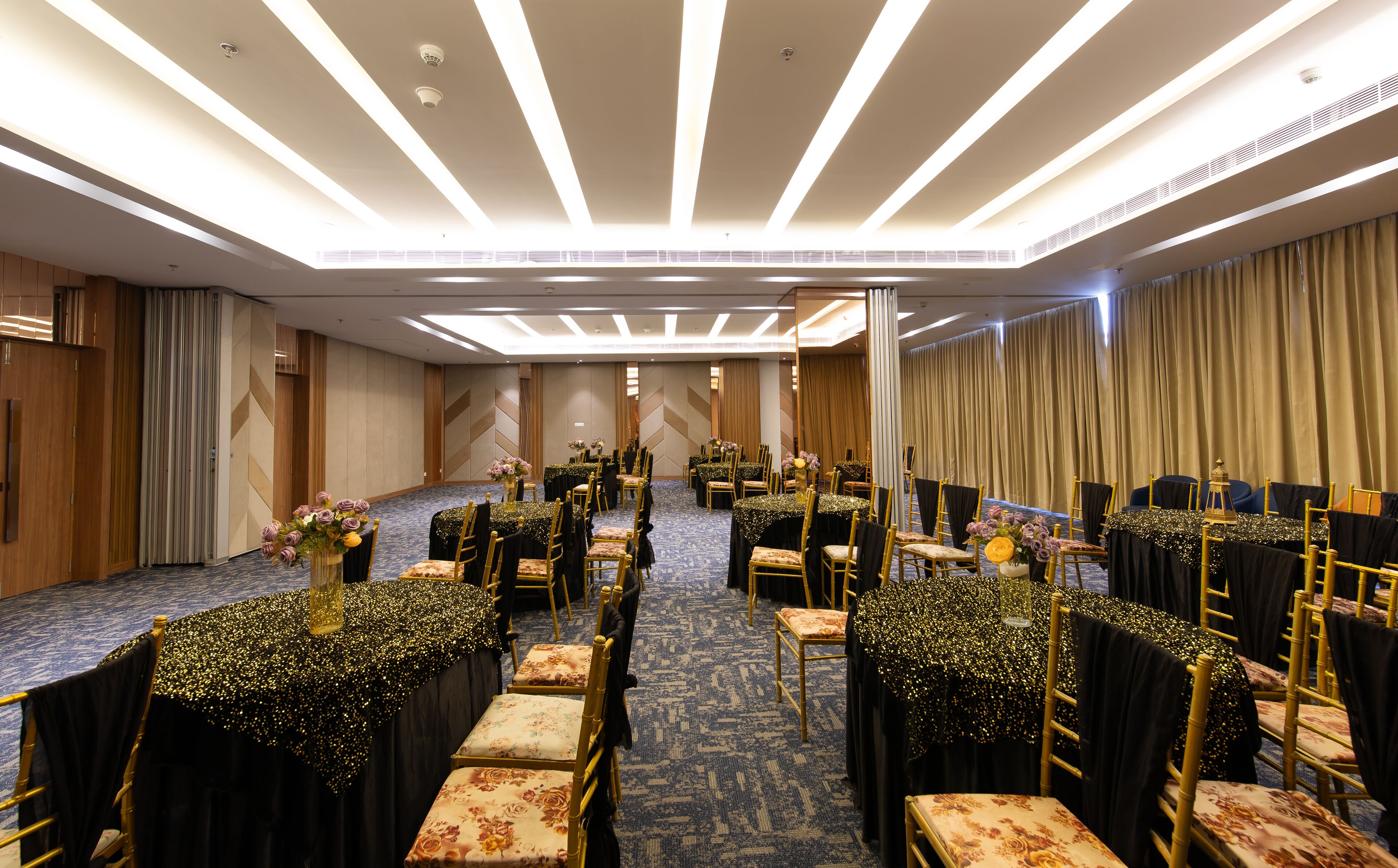Un salón de conferencias con filas de mesas y sillas, equipado con luces en el techo y suelo alfombrado.