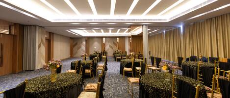 Un salón de conferencias con filas de mesas y sillas, equipado con luces en el techo y suelo alfombrado.