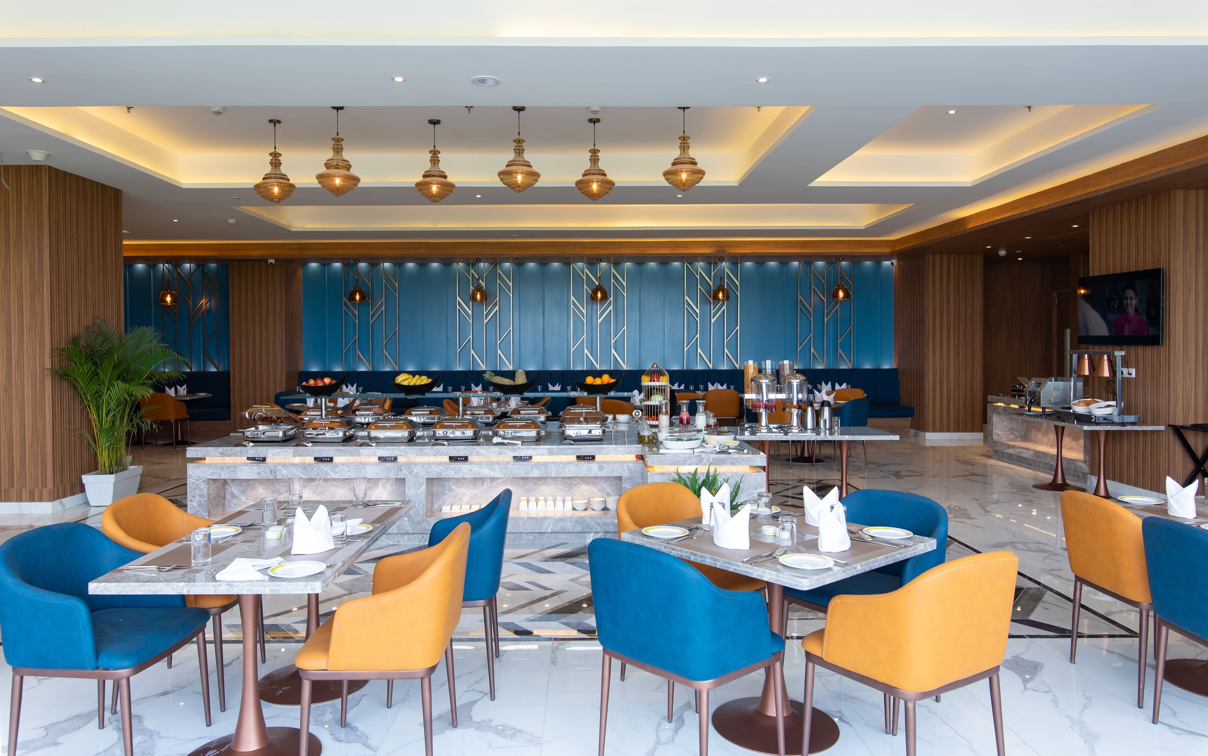 A modern restaurant with marble flooring, blue and orange chairs, a buffet with various food items, and pendant lighting.