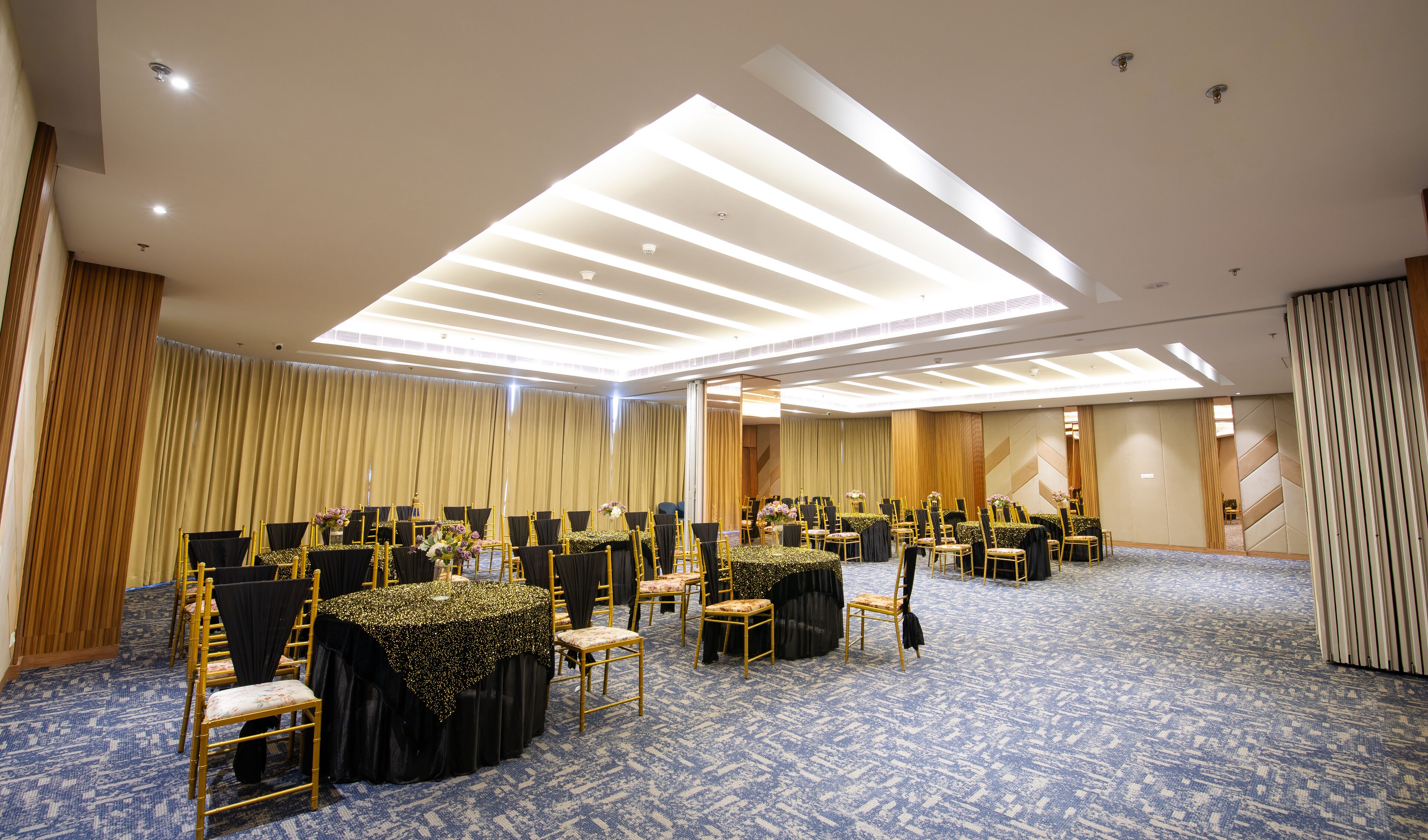 Un salón de conferencias con filas de mesas y sillas, una alfombra estampada y un techo con luminarias lineales.
