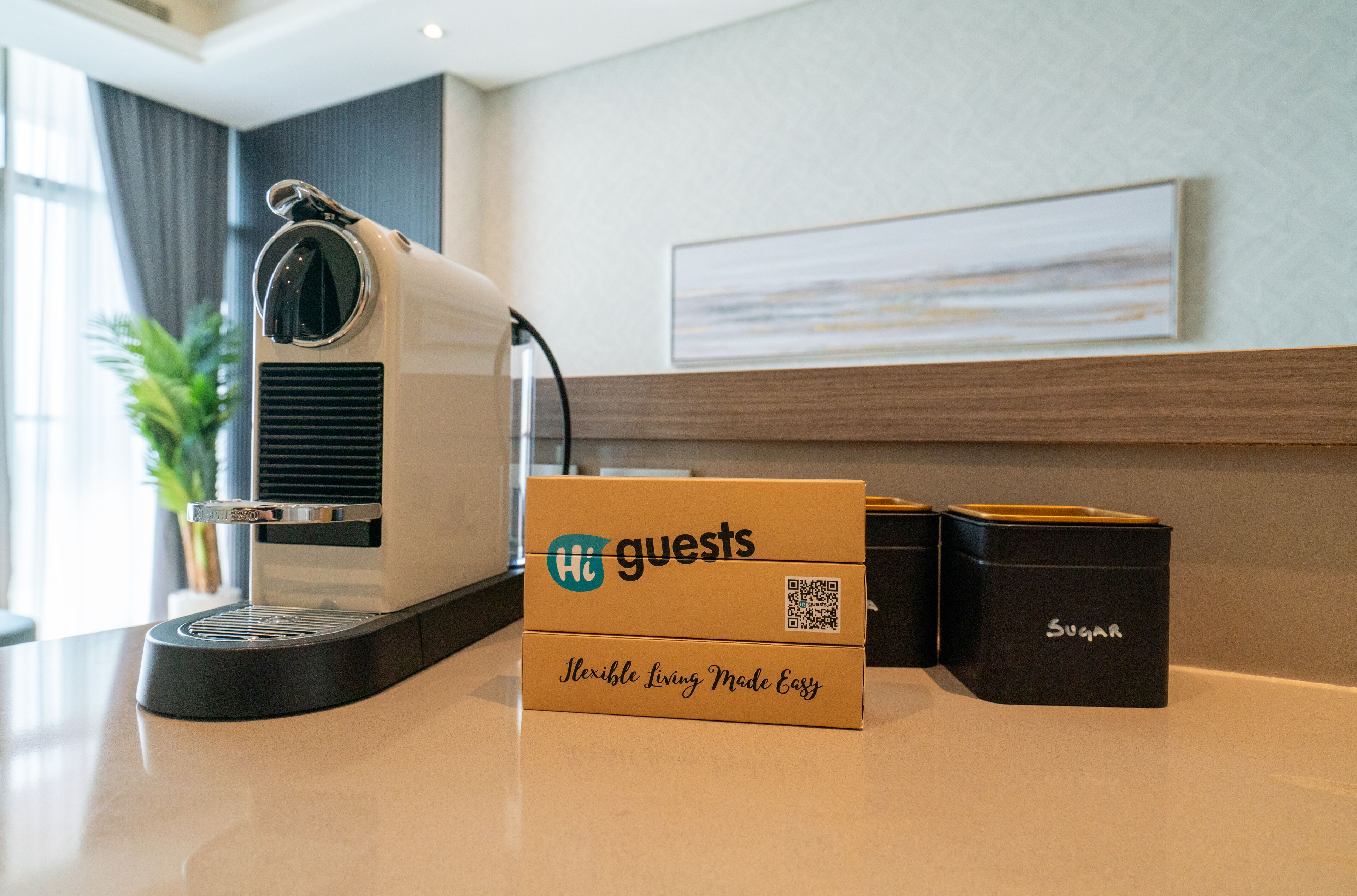 Uma máquina de café, uma caixa com a etiqueta 'Hi Guests' e dois recipientes em uma bancada.