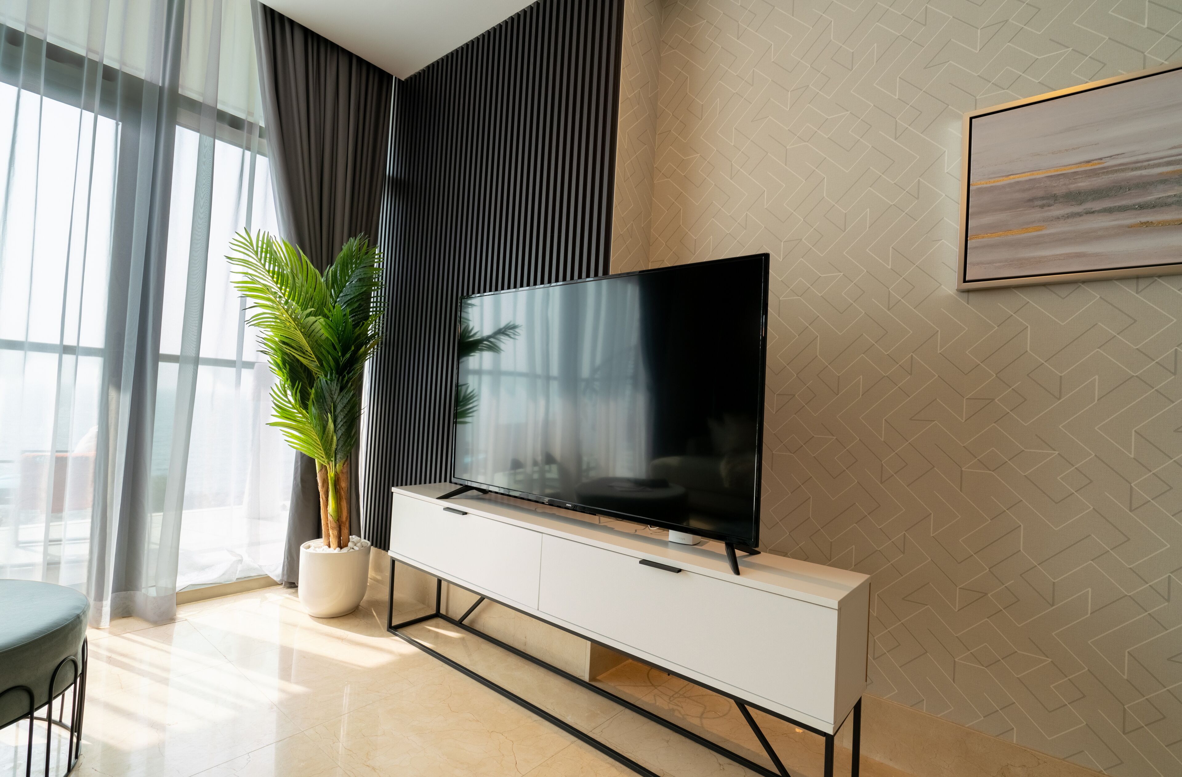 Un salon moderne avec un téléviseur à écran plat posé sur une console blanche, une plante en pot et une grande fenêtre ornée de rideaux légers.