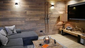 A modern living room with a wooden wall, a grey sofa, a coffee table with a book, a candle, and a small deer figurine. - Bergchalet Four Seasons (Reit im Winkl)