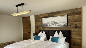 A modern bedroom with a large bed, wooden panel wall, a mountain painting, and a hanging light fixture. - Bergchalet Four Seasons (Reit im Winkl)