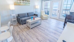 A modern living room with a grey sofa, a white coffee table, and a white armchair. There is a glass table with chairs outside a door leading to a balcony with a view of the beach. - Beachsid Bungalow @ Victorian Sleeps 8 (Galveston)