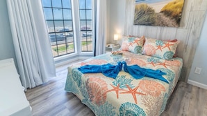 A bedroom with a bed, a window with curtains, a nightstand, and a painting on the wall. - Beachsid Bungalow @ Victorian Sleeps 8 (Galveston)