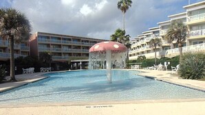 A swimming pool with a mushroom-shaped fountain, surrounded by palm trees and hotel buildings. - Beachsid Bungalow @ Victorian Sleeps 8 (Galveston)