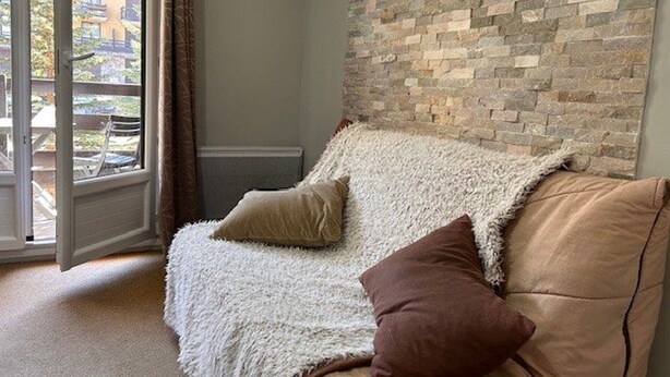 A cozy living room with a beige sofa, a fluffy white throw blanket, and two pillows. A large window with curtains offers a view of a building outside. - Cozy Studio for 4 with Ski Lift Access and South-Facing Balcony (Risoul)