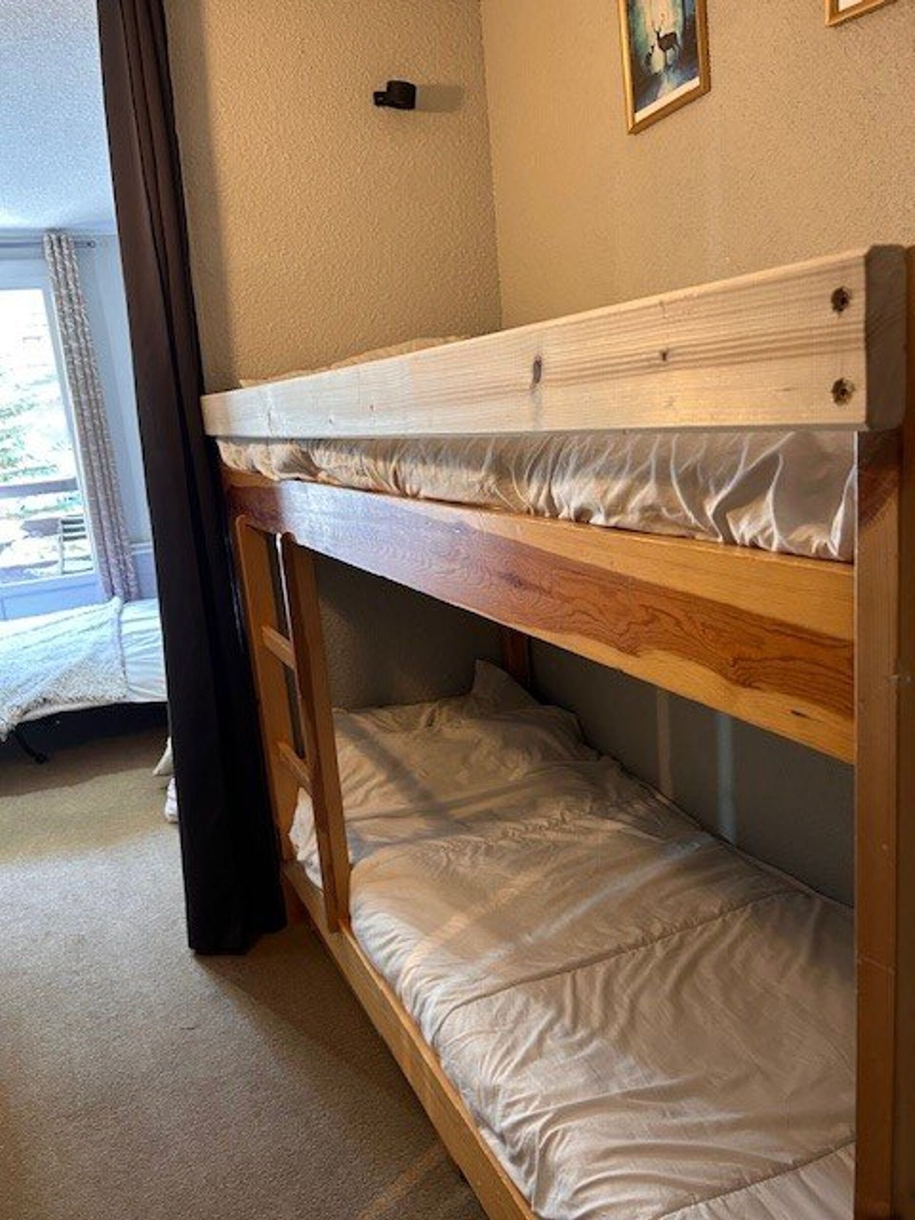 Ein Stockbett mit Matratze und Holzrahmen in einem Zimmer mit Teppichboden und einem Vorhangfenster.