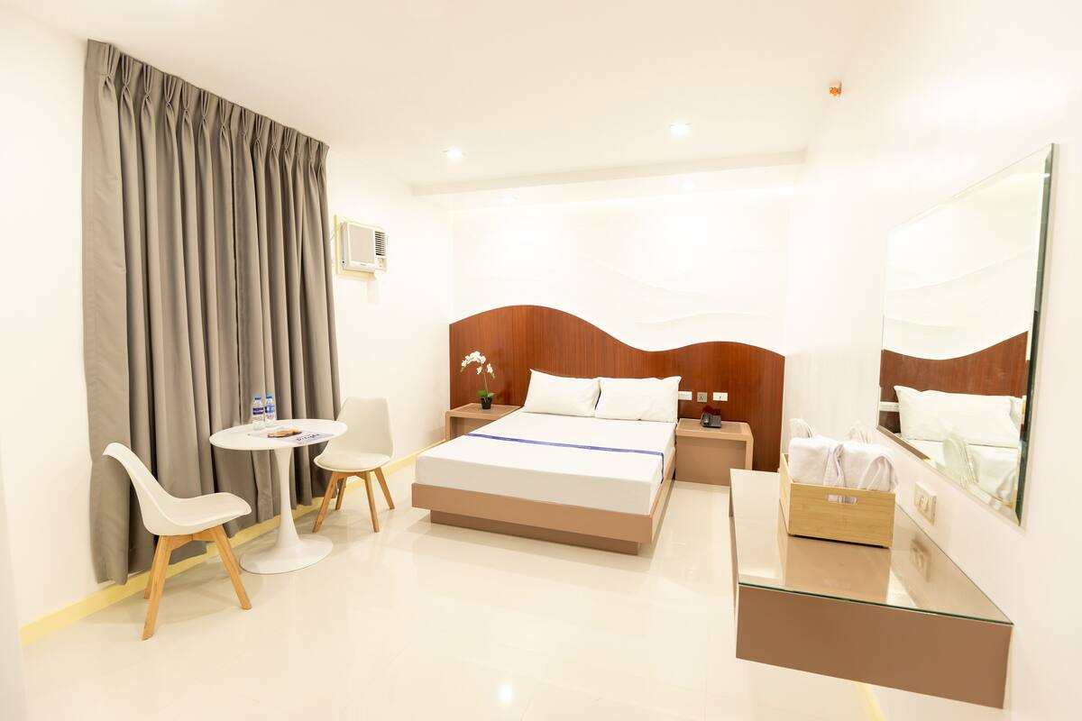 A modern hotel room with a large bed, a bedside table, a chair, a small table, and a mirror.