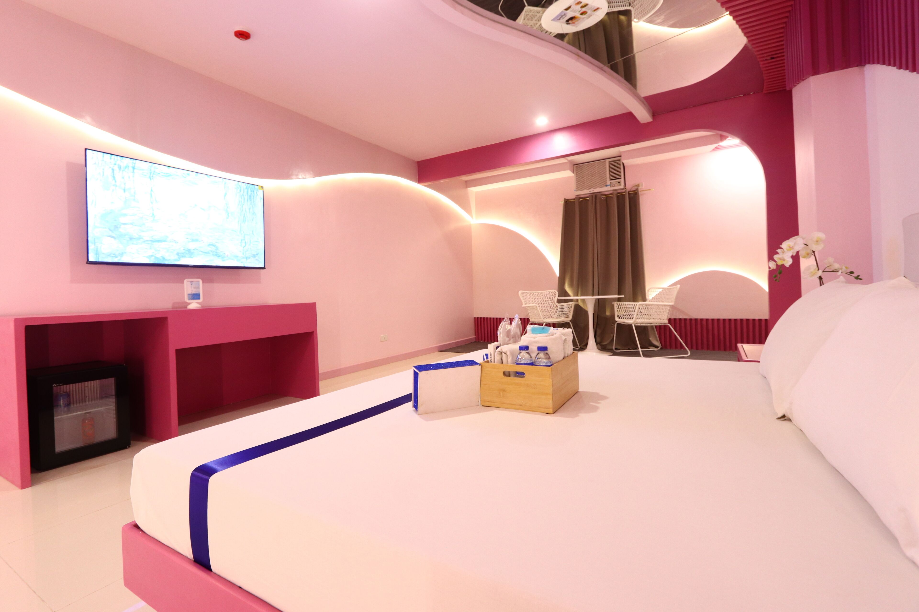 Ein modernes Hotelzimmer mit einem großen Bett, einem flachbildfernseher an der Wand, einer kleinen Sitzecke mit Sessel und Tisch sowie einem Holznachttisch mit einer Schachtel und Flaschen.