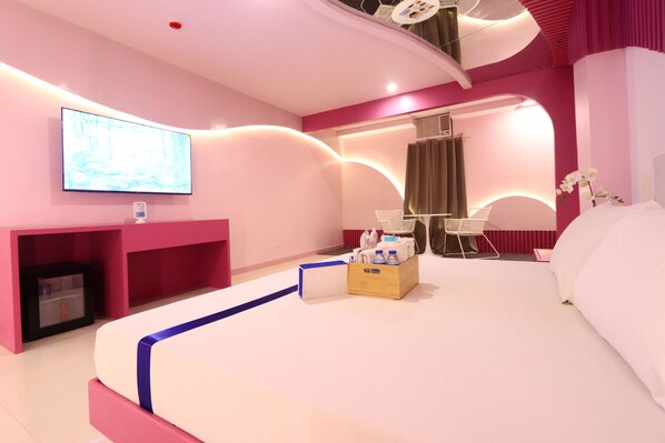 A modern hotel room with a large bed, a flat-screen TV mounted on the wall, a small seating area with a chair and table, and a wooden side table with a box and bottles. - Hotel Dreamworld East Ortigas (Cainta)