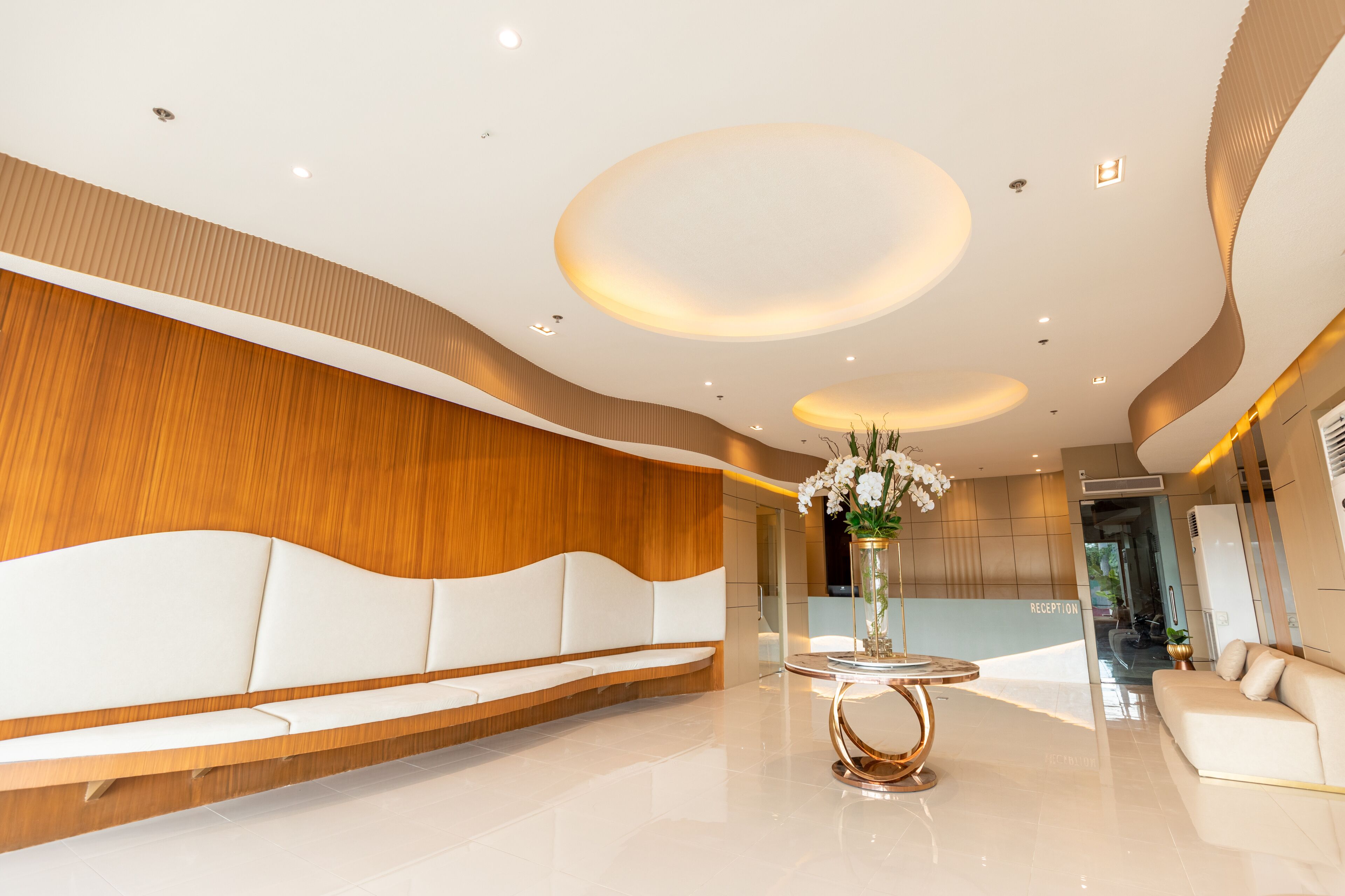 A modern reception area with a curved wooden wall, a white sofa, a round table with a vase of flowers, and a reception desk.