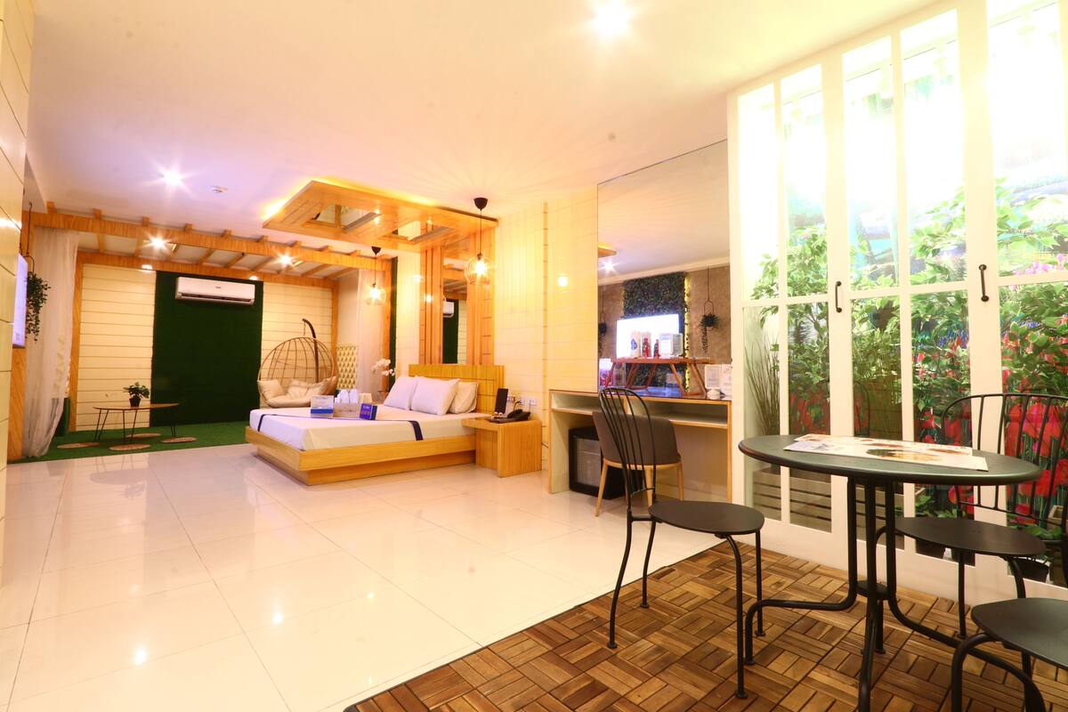 A modern hotel room with a bed, a desk, and a chair. There is a window with a view of plants.