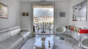 Una sala de estar moderna con un sofá blanco, una mesa de centro con figuritas, un comedor con mesa y sillas, y un balcón con vistas a edificios.