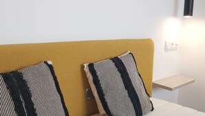 A room with a mustard-colored headboard, two striped pillows, a small wooden shelf, and a pendant light.