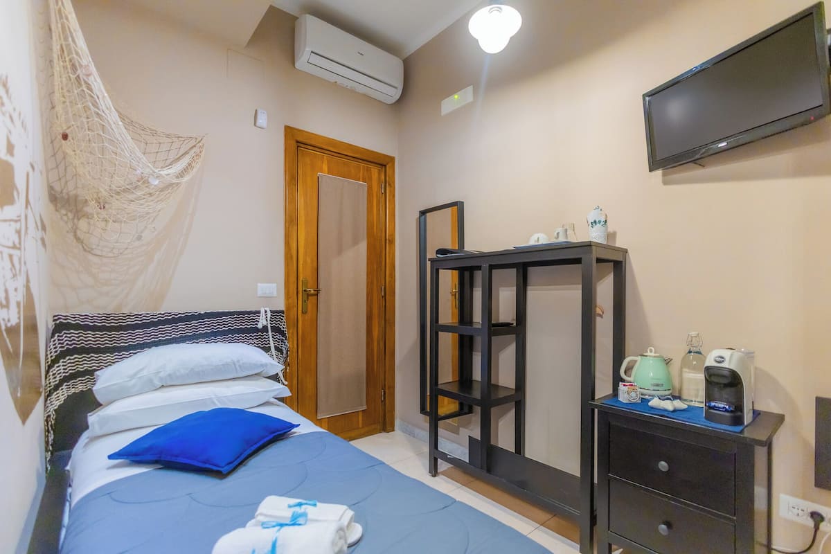 A bedroom with a bed, a bedside table with a kettle, a coffee maker, and a TV on the wall.