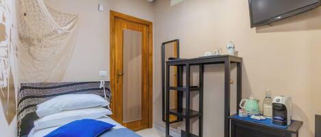 A bedroom with a bed, a bedside table with a kettle, a coffee maker, and a TV on the wall.